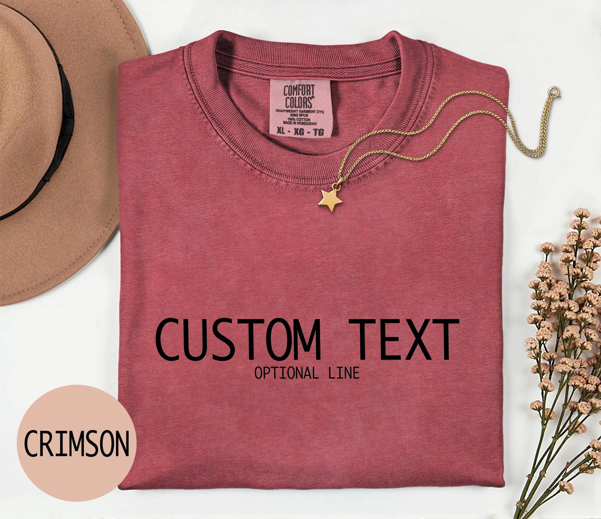 a red t-shirt with the text &amp;quot;CUSTOM TEXT&amp;quot; printed on it, along with the word &amp;quot;CRIMSON&amp;quot; in a smaller font. The t-shirt is laid out on a white surface, accompanied by a hat, a necklace, and some flowers.