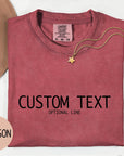 a red t-shirt with the text "CUSTOM TEXT" printed on it, along with the word "CRIMSON" in a smaller font. The t-shirt is laid out on a white surface, accompanied by a hat, a necklace, and some flowers.