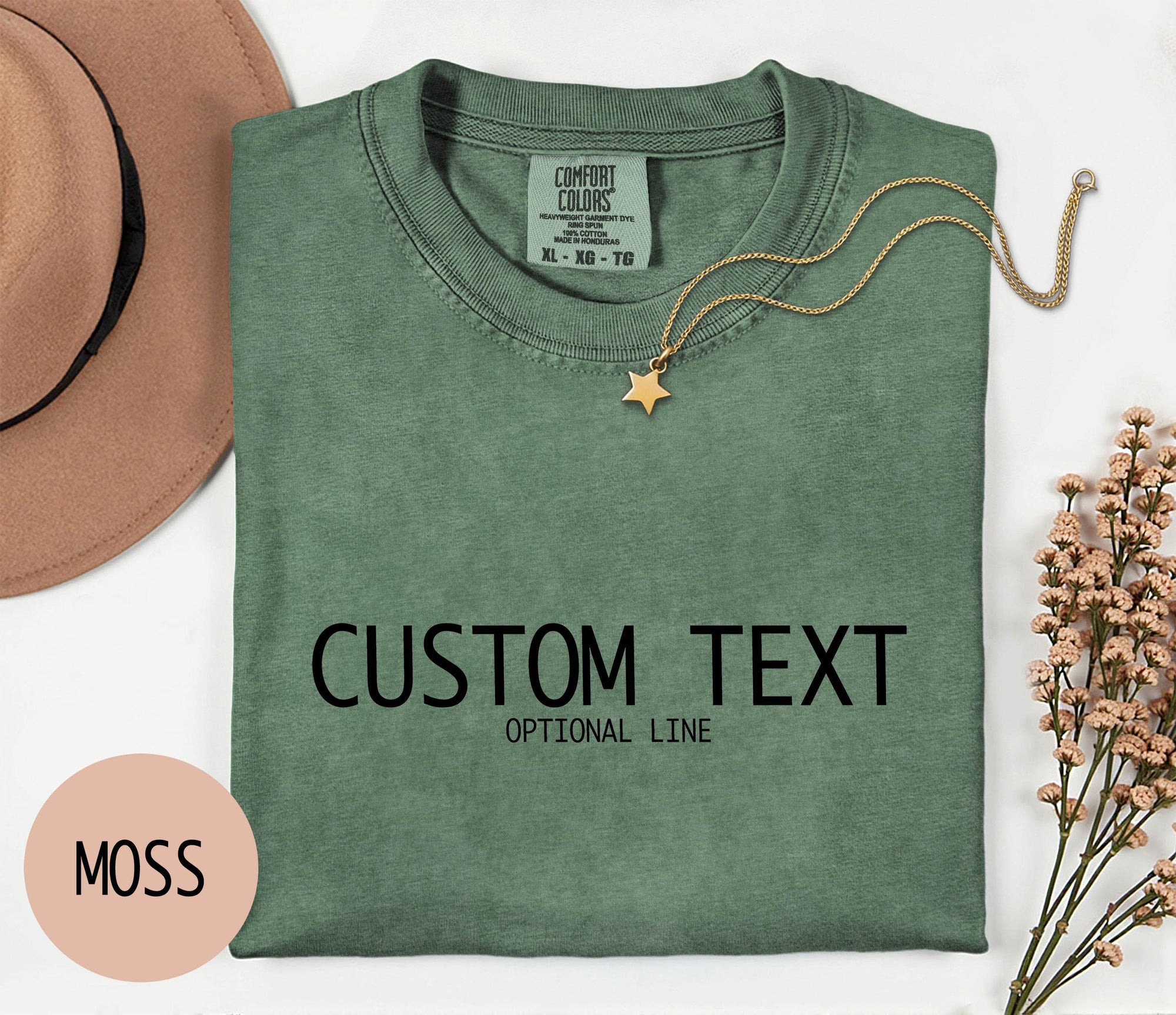 a green t-shirt with the text &amp;quot;CUSTOM TEXT&amp;quot; printed on it, along with the phrase &amp;quot;OPTIONAL LINE&amp;quot; below it. The t-shirt is laid out on a white surface, accompanied by a hat, a necklace, and some flowers.