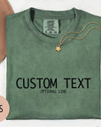 a green t-shirt with the text "CUSTOM TEXT" printed on it, along with the phrase "OPTIONAL LINE" below it. The t-shirt is laid out on a white surface, accompanied by a hat, a necklace, and some flowers.