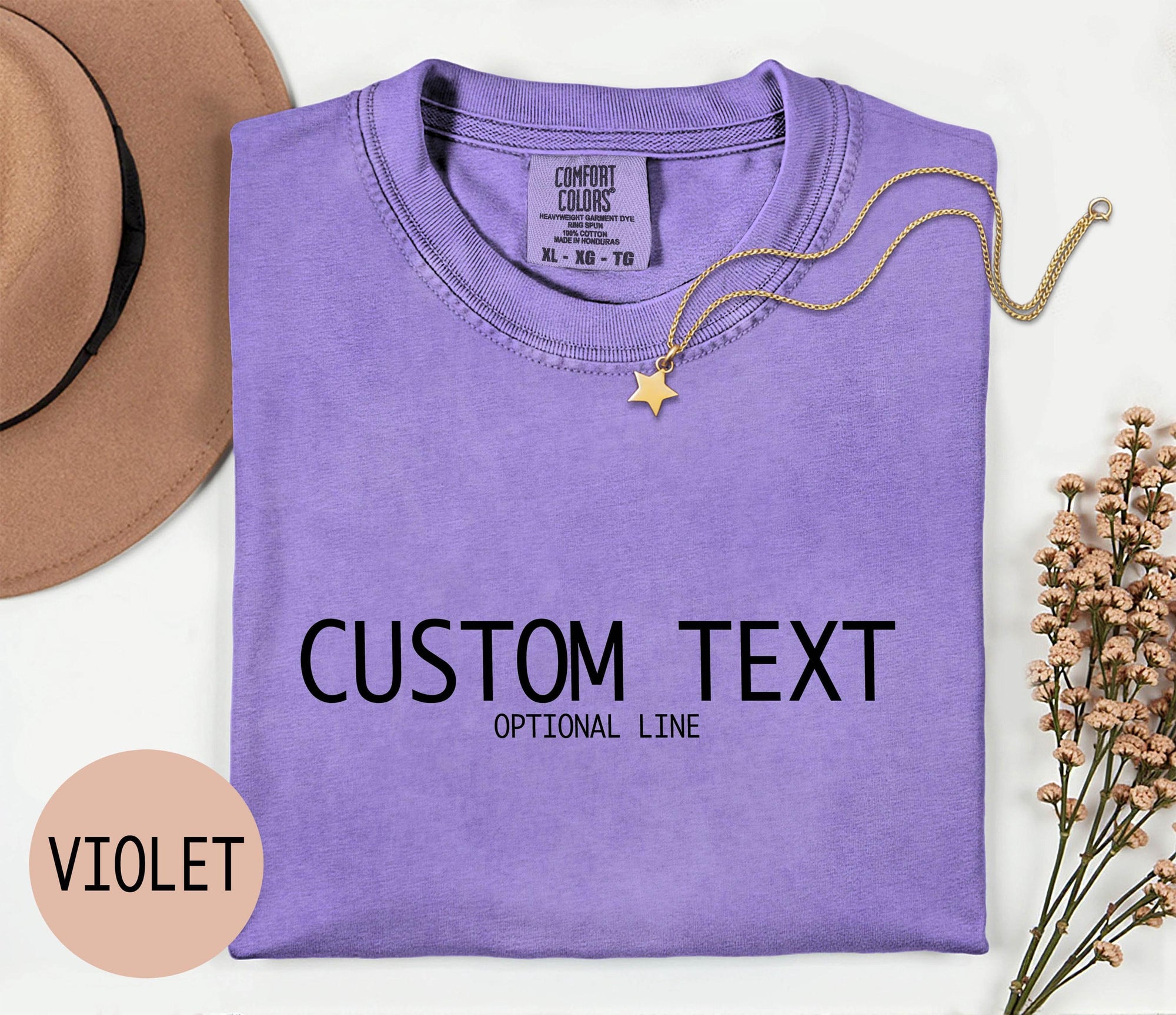 a purple t-shirt with the text &amp;quot;CUSTOM TEXT&amp;quot; and &amp;quot;OPTIONAL LINE&amp;quot; printed on it, along with a gold star charm and a beige hat. The t-shirt is laid out on a white surface, and there is a small pink circle with the word &amp;quot;VIOLET&amp;quot; written on it.