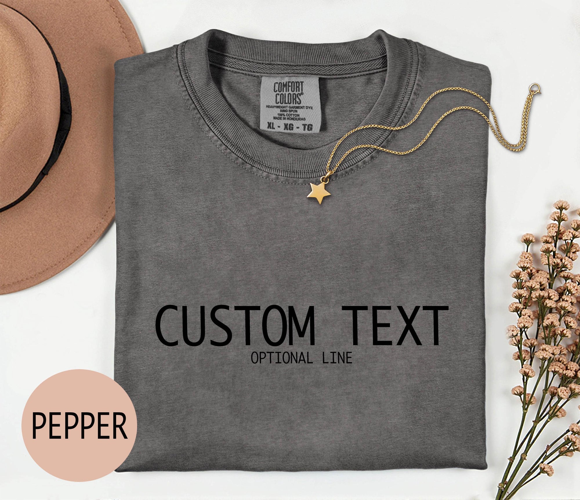 a gray t-shirt with the text &amp;quot;CUSTOM TEXT&amp;quot; printed on it, along with the word &amp;quot;PEPPER&amp;quot; in a smaller font. The t-shirt is laid out on a white surface, accompanied by a hat, a necklace, and some flowers.