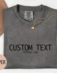 a gray t-shirt with the text "CUSTOM TEXT" printed on it, along with the word "PEPPER" in a smaller font. The t-shirt is laid out on a white surface, accompanied by a hat, a necklace, and some flowers.