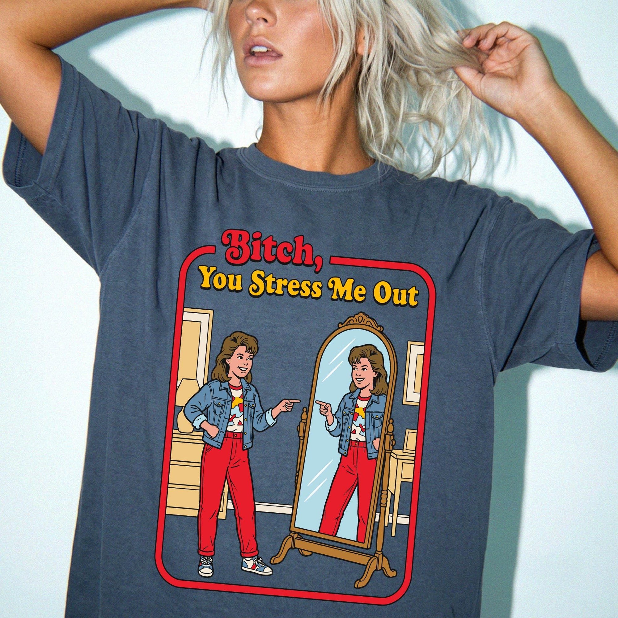 A woman with blonde hair wearing a blue t-shirt with a graphic design depicting a woman looking into a mirror and pointing at herself.