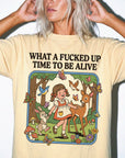 A woman wearing a yellow t-shirt with a graphic design depicting a young girl surrounded by various animals, including a deer, rabbit, and butterflies, along with the text "What a FUCKED UP TIME TO BE ALIVE".