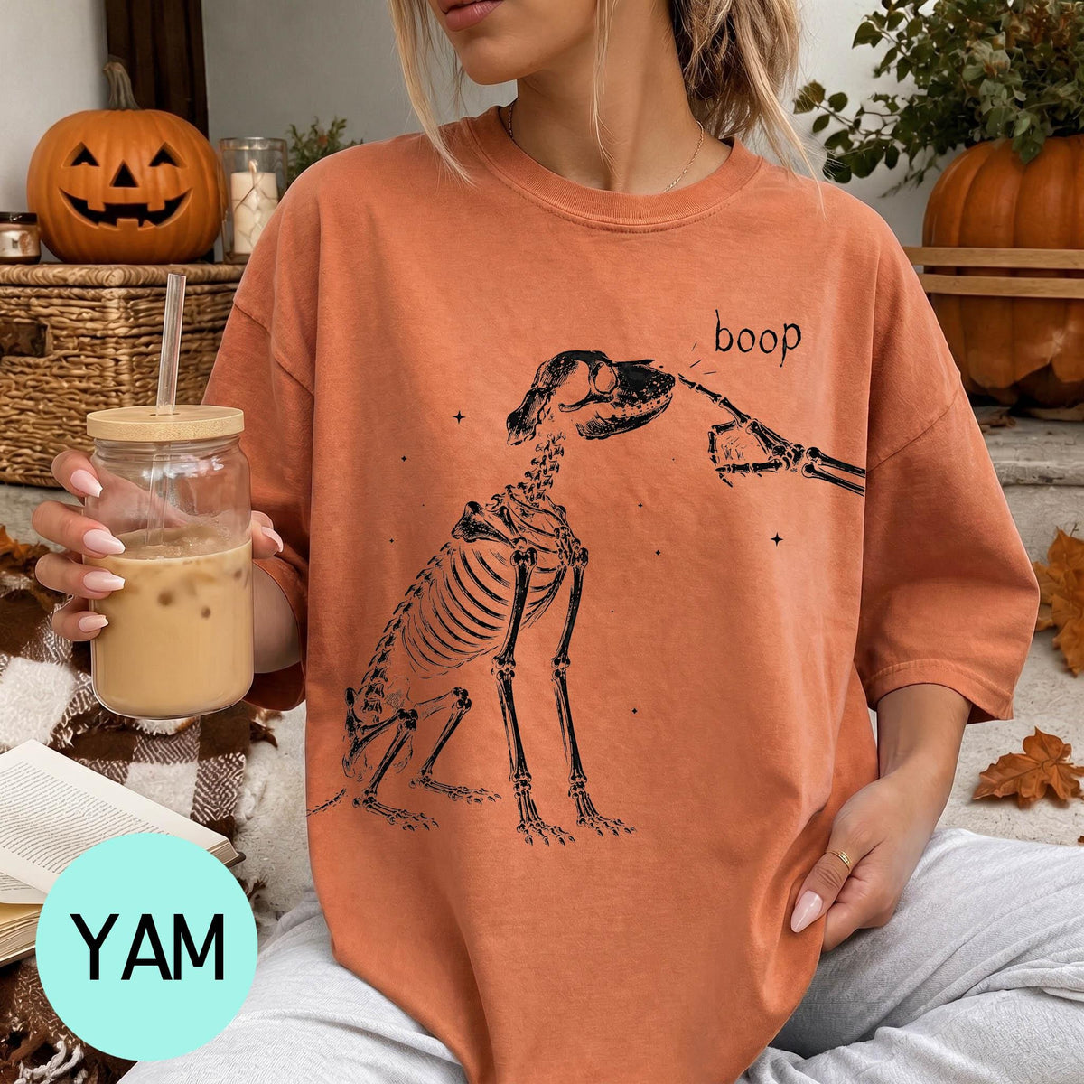 A woman wearing an orange t-shirt with a skeleton drawing and the word "boop" printed on it, holding a glass of iced coffee.