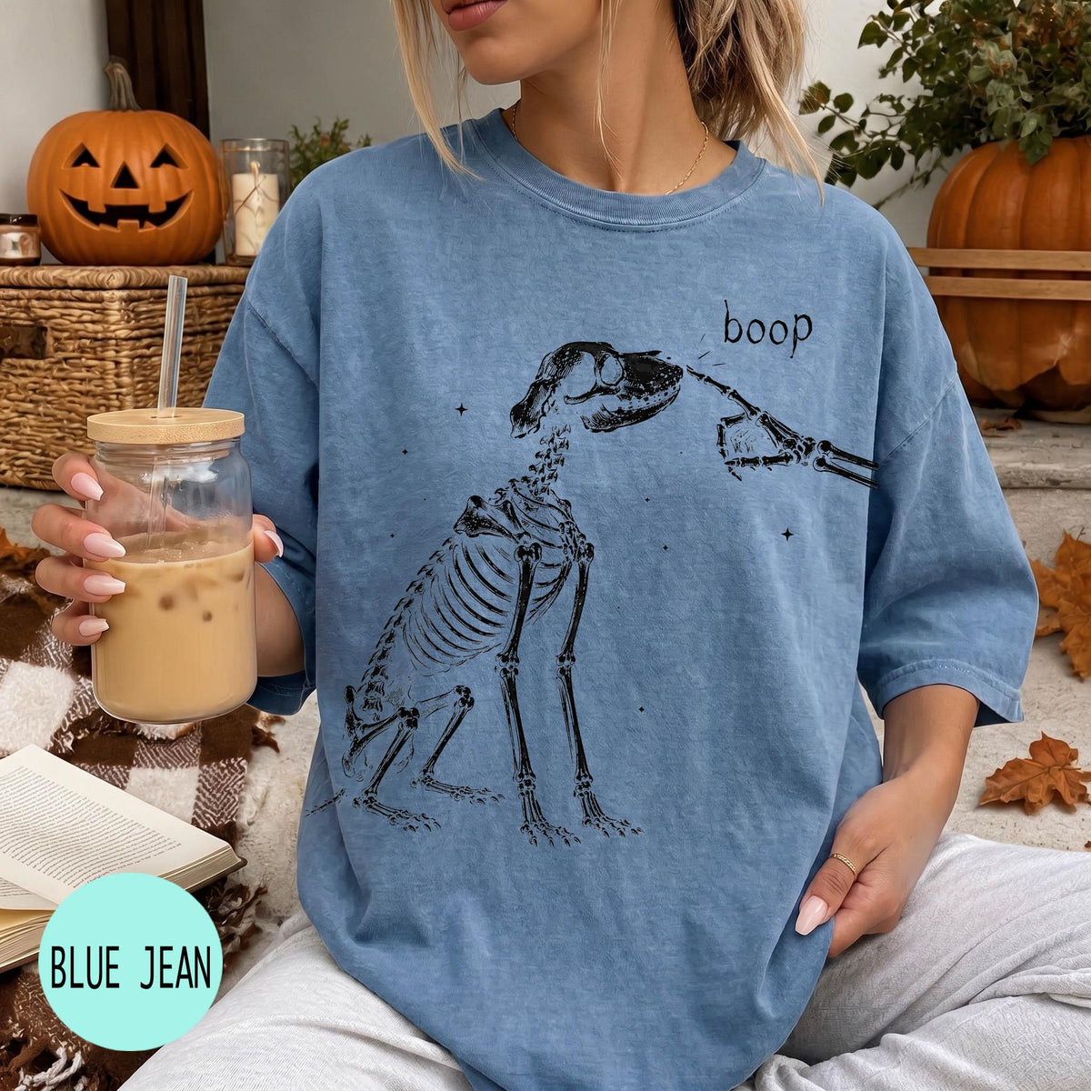 A woman wearing a blue t-shirt with a skeleton drawing and the word "boop" printed on it, holding a glass of iced coffee.