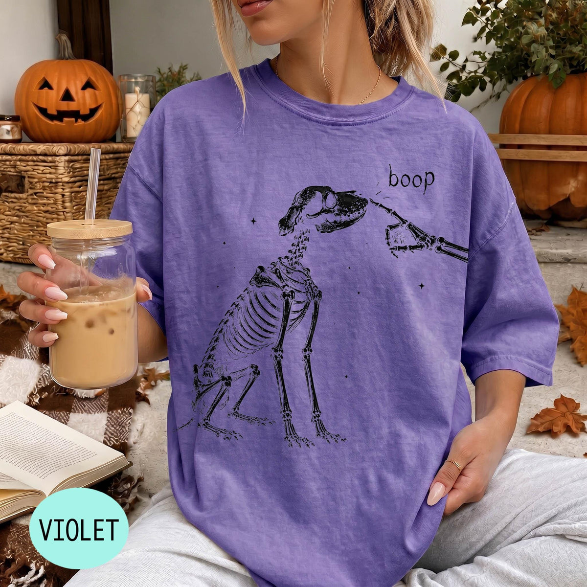 A woman wearing a purple t-shirt with a skeleton drawing and the word &amp;quot;boop&amp;quot; printed on it, holding a glass of iced coffee.