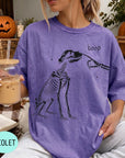 A woman wearing a purple t-shirt with a skeleton drawing and the word "boop" printed on it, holding a glass of iced coffee.