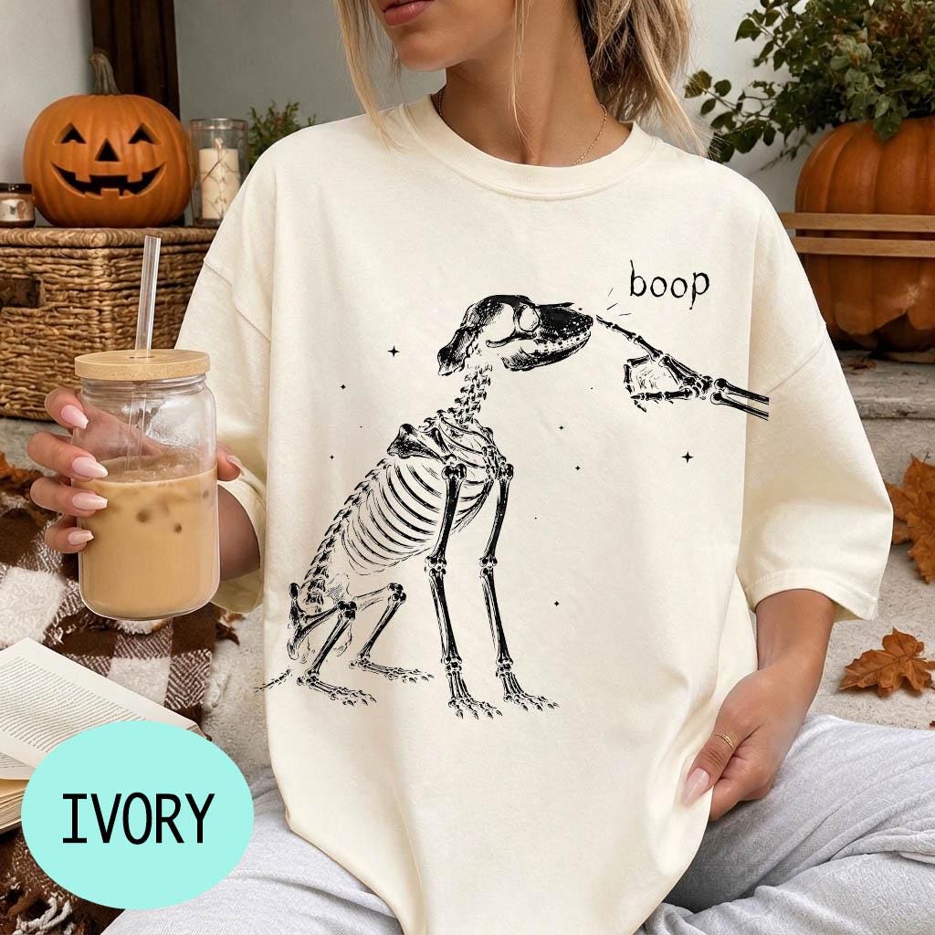 A woman wearing a white t-shirt with a skeleton graphic and the word &amp;quot;boop&amp;quot; printed on it, holding a glass of iced coffee.