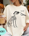 A woman wearing a white t-shirt with a skeleton graphic and the word "boop" printed on it, holding a glass of iced coffee.