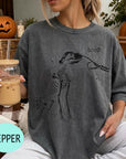 A woman wearing a gray t-shirt with a skeleton drawing and the word "boop" printed on it, holding a glass of iced coffee.