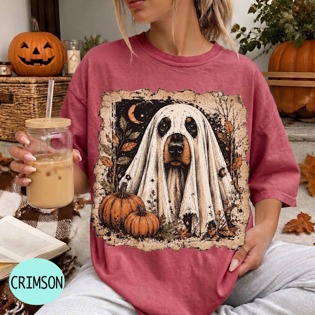 A woman wearing a pink t-shirt with a ghost design and holding a glass of iced coffee, with a pumpkin and book visible in the background.