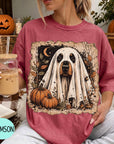 A woman wearing a pink t-shirt with a ghost design and holding a glass of iced coffee, with a pumpkin and book visible in the background.