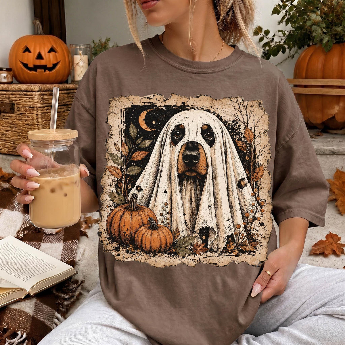 A woman is sitting on a couch, wearing a t-shirt with a Halloween-themed design featuring a ghost and pumpkins. She is holding a glass of iced coffee and has a book nearby.