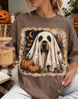 A woman is sitting on a couch, wearing a t-shirt with a Halloween-themed design featuring a ghost and pumpkins. She is holding a glass of iced coffee and has a book nearby.