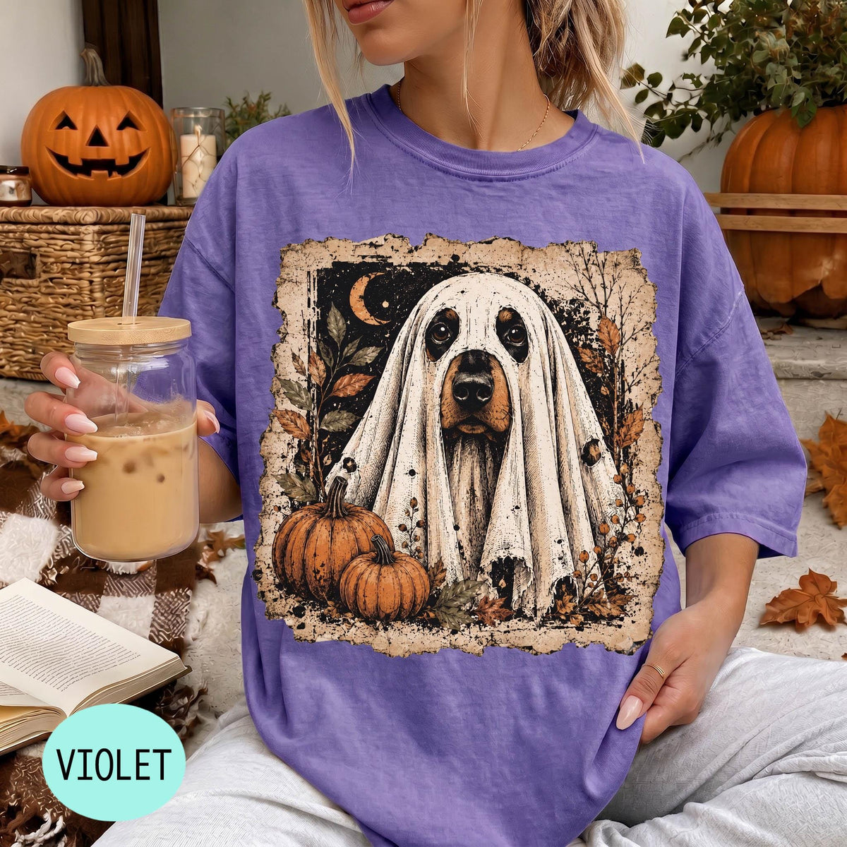 A woman wearing a purple t-shirt with a graphic of a ghost dog holding a pumpkin and a glass of iced coffee.