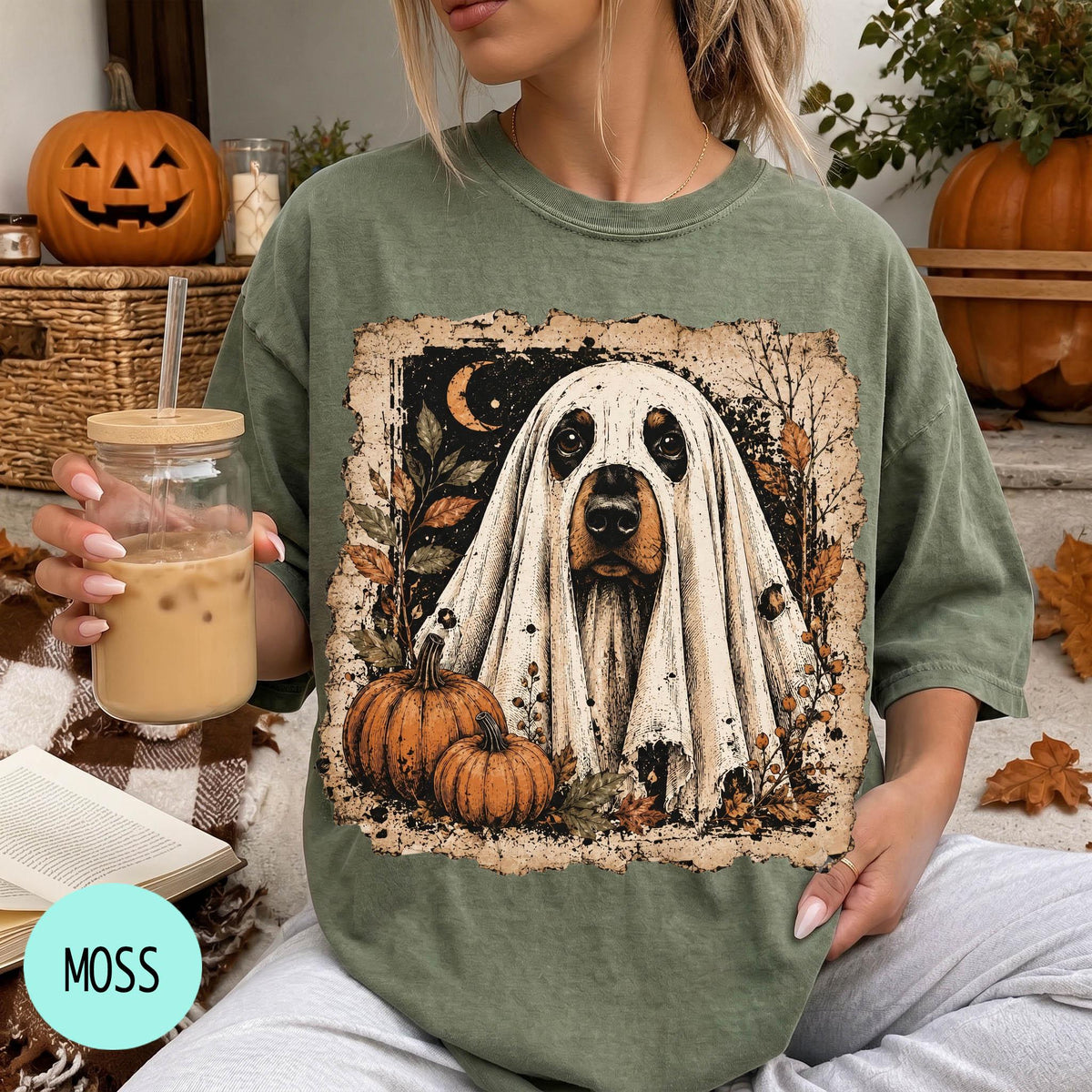A woman is sitting on a couch, wearing a green t-shirt with a graphic of a ghost dog and pumpkins. She is holding a glass of iced coffee.