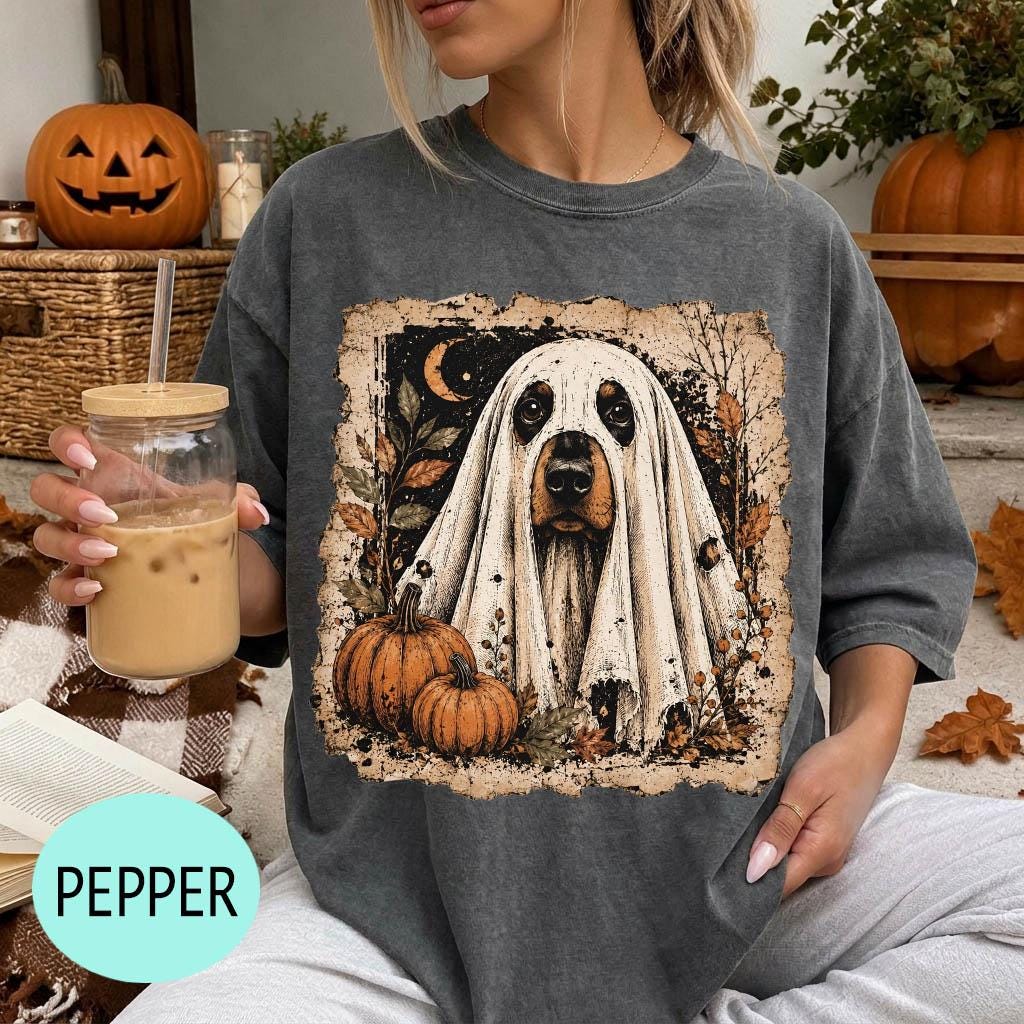 A woman is sitting on a couch, wearing a gray t-shirt with a Halloween-themed graphic of a ghost dog surrounded by pumpkins and a jack-o-lantern. She is holding a glass of iced coffee.