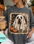 A woman is sitting on a couch, wearing a gray t-shirt with a Halloween-themed graphic of a ghost dog surrounded by pumpkins and a jack-o-lantern. She is holding a glass of iced coffee.