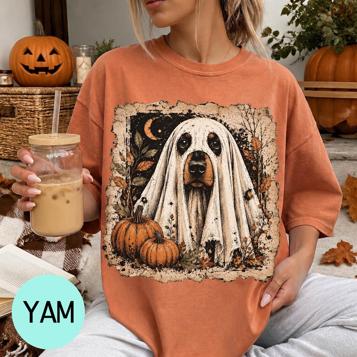 A woman is sitting on a couch, wearing a t-shirt with a Halloween-themed graphic of a ghost dog. She is holding a drink in a glass with a straw.