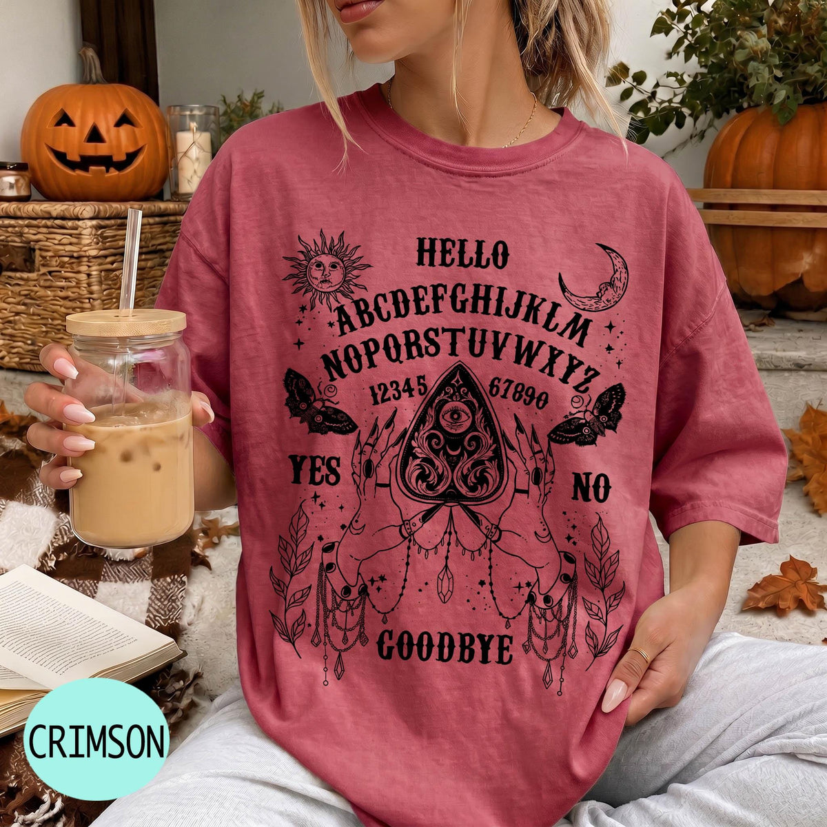 A woman wearing a red t-shirt with a Ouija board design and text printed on it, holding a drink in a glass.