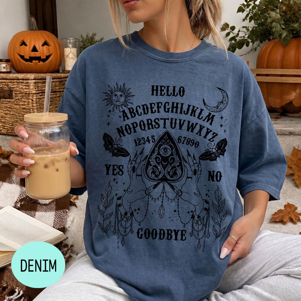 A woman wearing a blue t-shirt with a Ouija board design and holding a drink is sitting in front of a Halloween decoration.