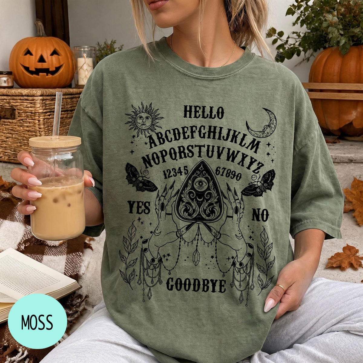 A woman wearing a green t-shirt with an Ouija board design and text printed on it, holding a drink in a glass.