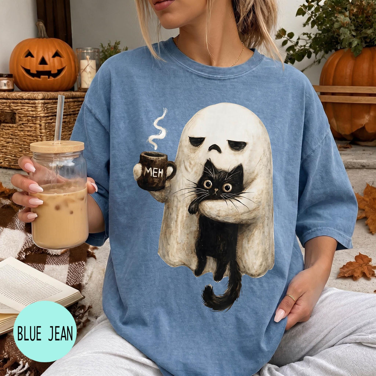 A woman wearing a blue t-shirt with a graphic of a ghost holding a coffee mug and a black cat, sitting on a couch with a pumpkin and a book nearby.