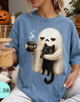 A woman wearing a blue t-shirt with a graphic of a ghost holding a coffee mug and a black cat, sitting on a couch with a pumpkin and a book nearby.