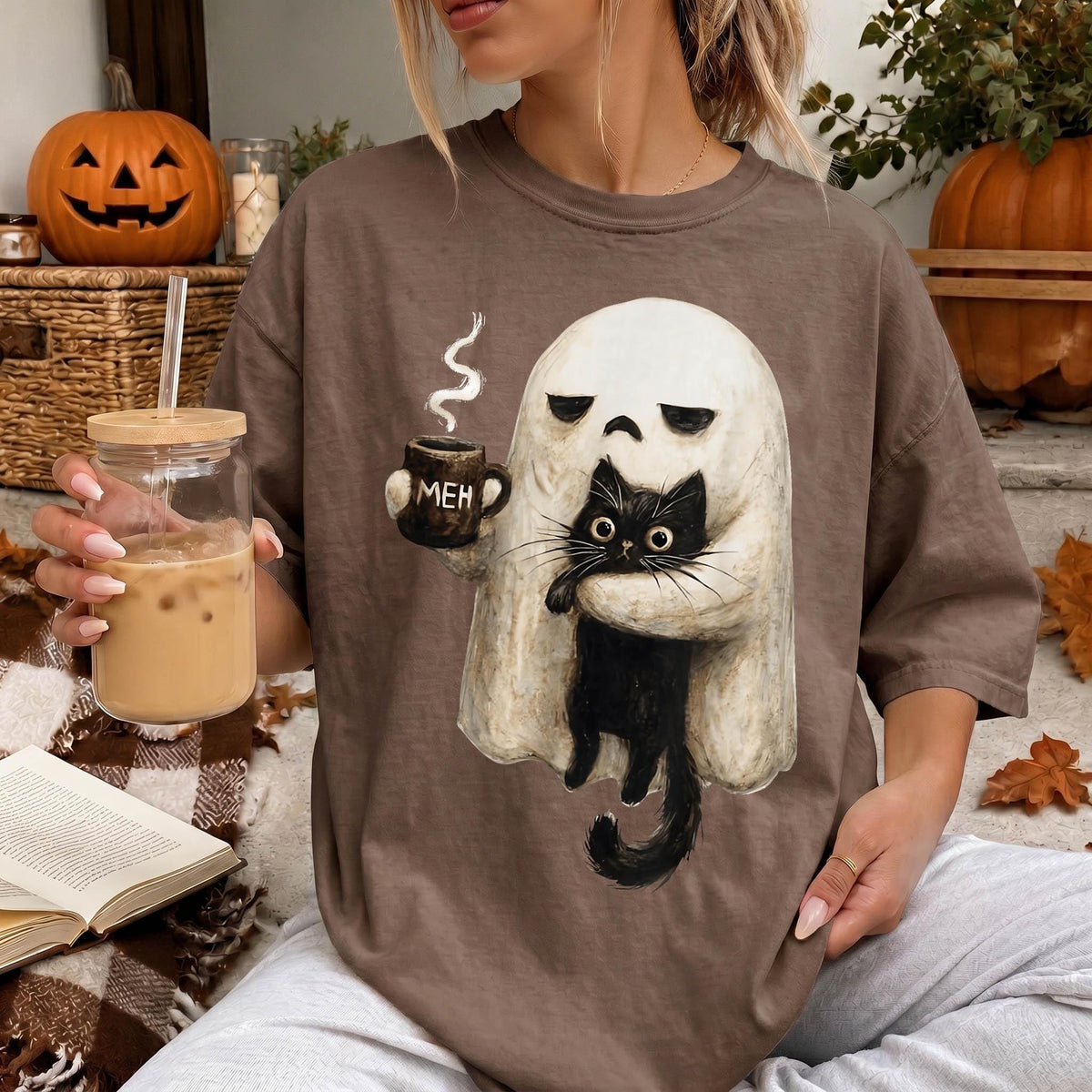 A woman is sitting on a couch, wearing a t-shirt with a graphic of a ghost holding a coffee mug and a black cat. She is holding a glass of iced coffee and a book.