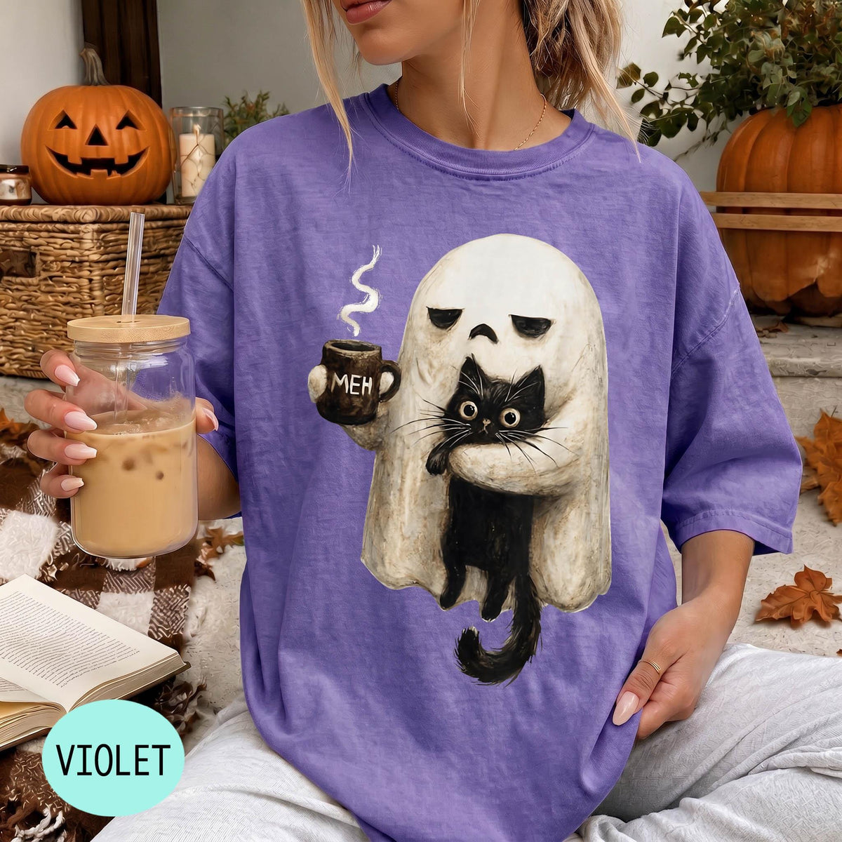 A person wearing a purple t-shirt with a graphic of a ghost holding a coffee mug and a black cat, sitting on a couch with a pumpkin and a book nearby.
