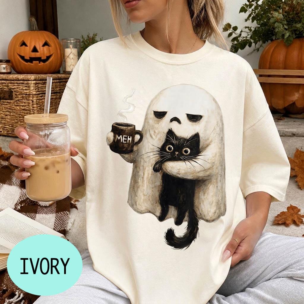 A person wearing a white t-shirt with a graphic of a ghost holding a coffee mug and a black cat, with a pumpkin and a book visible in the background.
