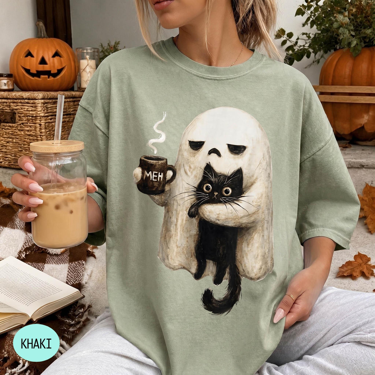 A woman wearing a green t-shirt with a graphic of a ghost holding a coffee mug and a cat, sitting in a cozy setting with a pumpkin and a book.