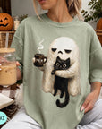 A woman wearing a green t-shirt with a graphic of a ghost holding a coffee mug and a cat, sitting in a cozy setting with a pumpkin and a book.