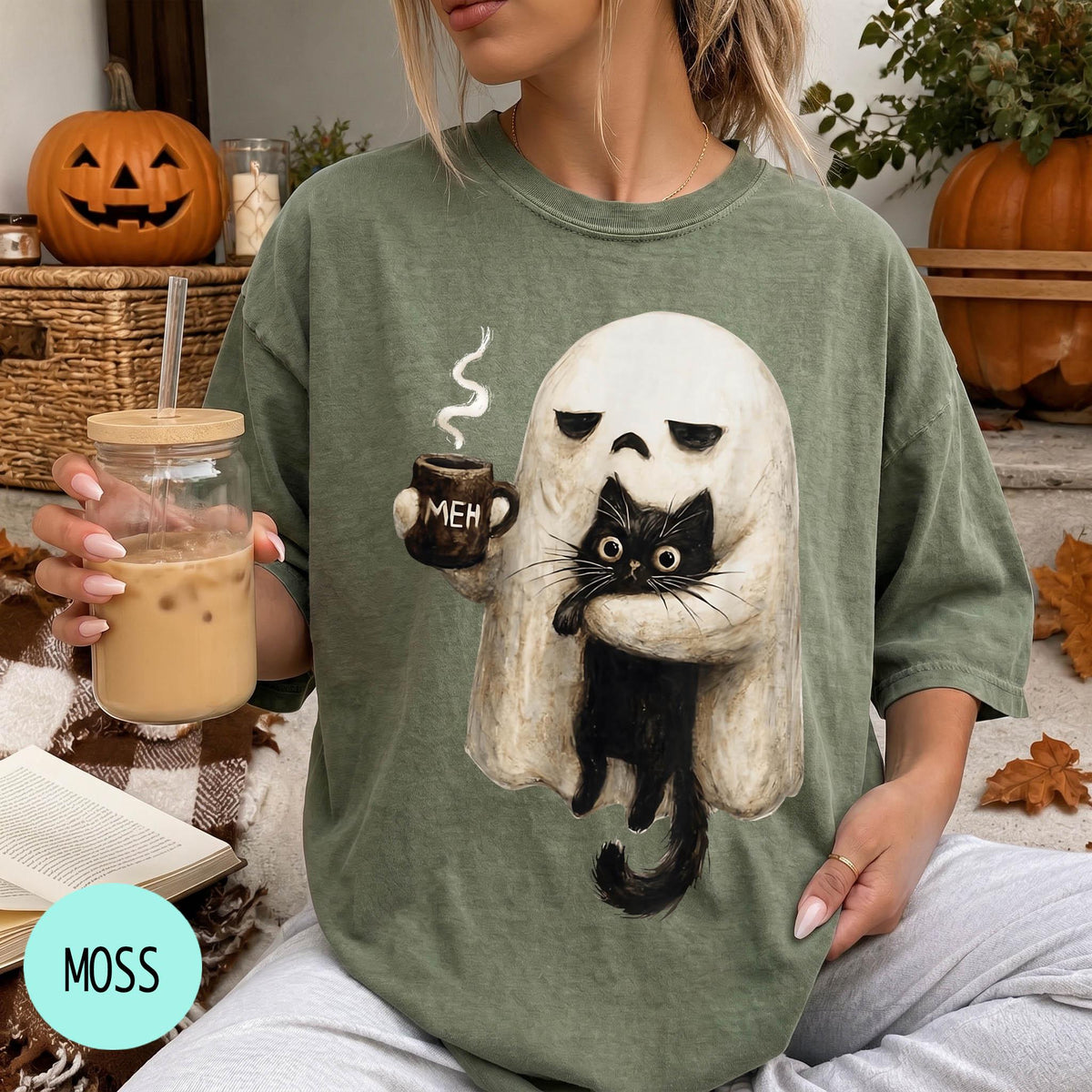 A person is sitting on a couch, wearing a green t-shirt with a graphic of a ghost holding a coffee mug and a cat.