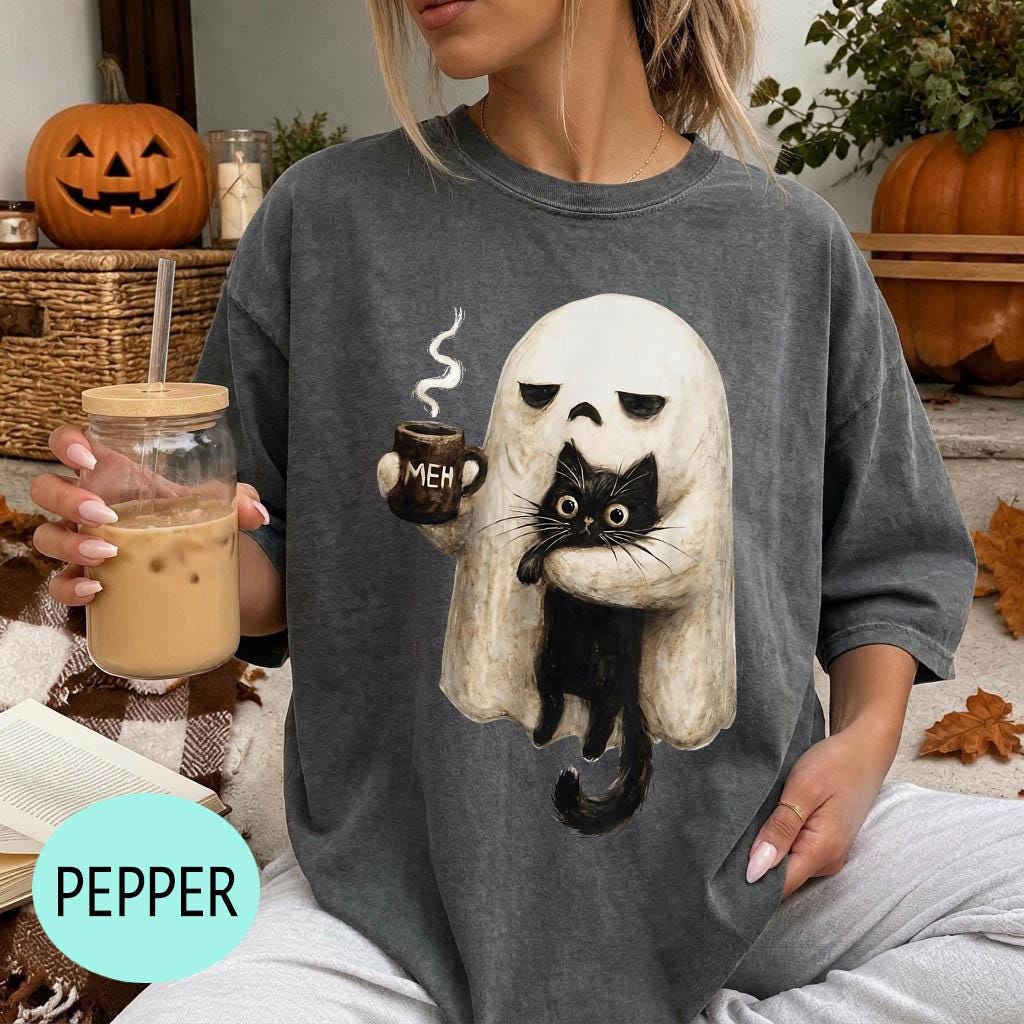 A woman wearing a gray t-shirt with a graphic of a ghost holding a coffee mug and a cat, sitting on a couch with a pumpkin and a book nearby.