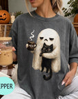 A woman wearing a gray t-shirt with a graphic of a ghost holding a coffee mug and a cat, sitting on a couch with a pumpkin and a book nearby.