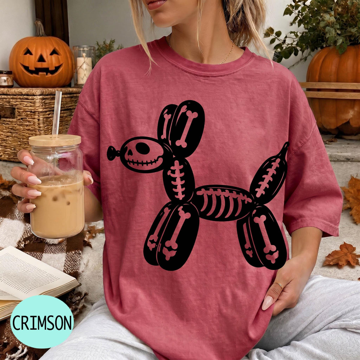A woman wearing a pink t-shirt with a skeleton dog design is sitting in a room with Halloween decorations, including a carved pumpkin and a skeleton decoration.