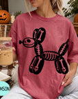 A woman wearing a pink t-shirt with a skeleton dog design is sitting in a room with Halloween decorations, including a carved pumpkin and a skeleton decoration.