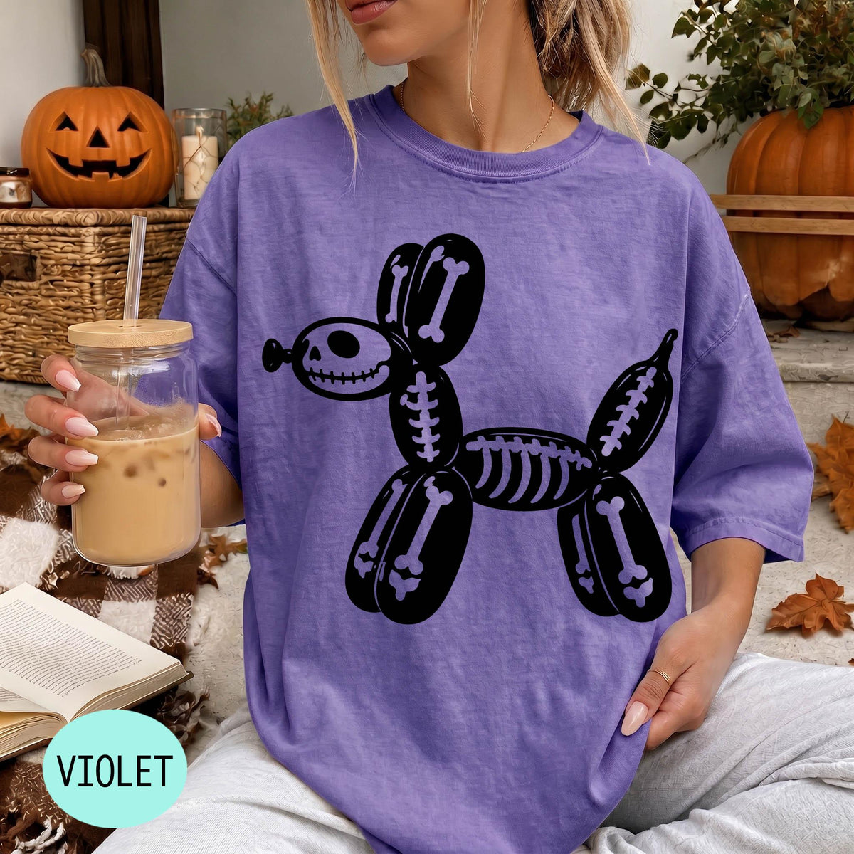 A woman wearing a purple t-shirt with a skeleton dog graphic is sitting on a blanket, holding a drink in a glass.