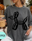 A woman wearing a gray t-shirt with a skeleton dog graphic is sitting on a couch, holding a drink in a glass.