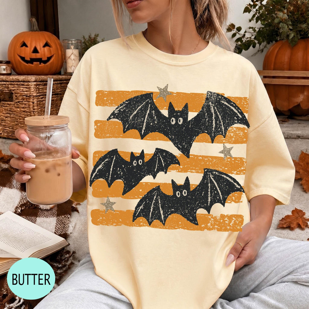 A woman wearing a yellow t-shirt with a graphic of bats and stars is sitting on a couch, holding a drink in a glass.