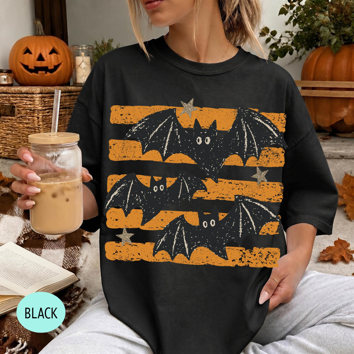 A woman wearing a black t-shirt with a graphic of bats and stars is sitting in a room with Halloween decorations, including a jack-o-lantern and a pumpkin. She is holding a drink in a glass with a straw.