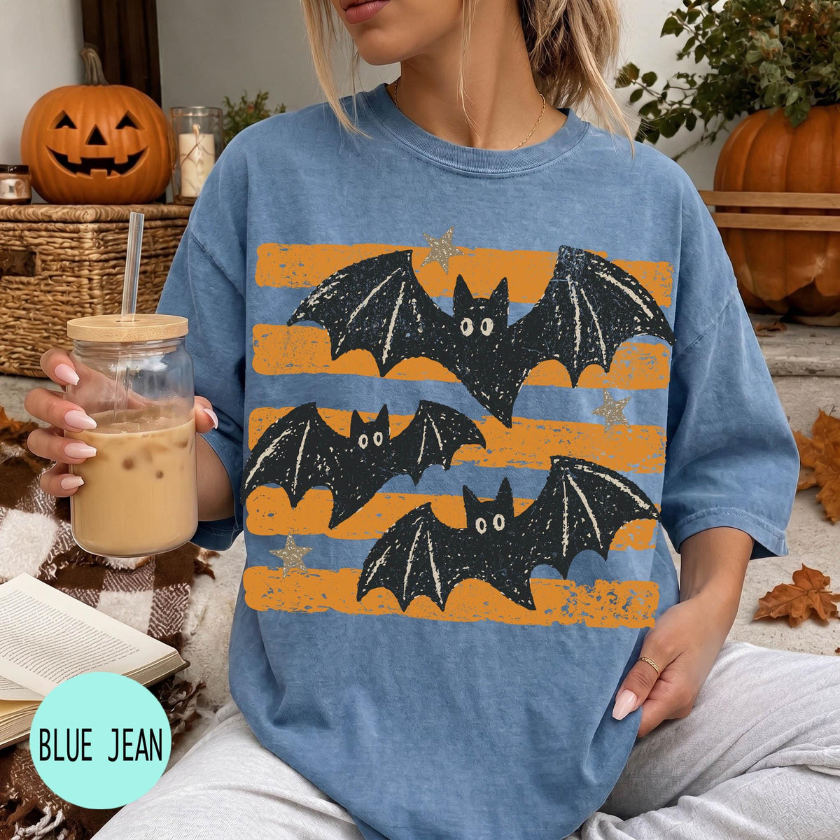 A woman wearing a blue t-shirt with a graphic of bats and orange and black stripes, holding a drink and sitting on a blanket with a pumpkin and other fall decorations in the background.
