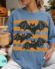 A woman wearing a blue t-shirt with a graphic of bats and orange and black stripes, holding a drink and sitting on a blanket with a pumpkin and other fall decorations in the background.