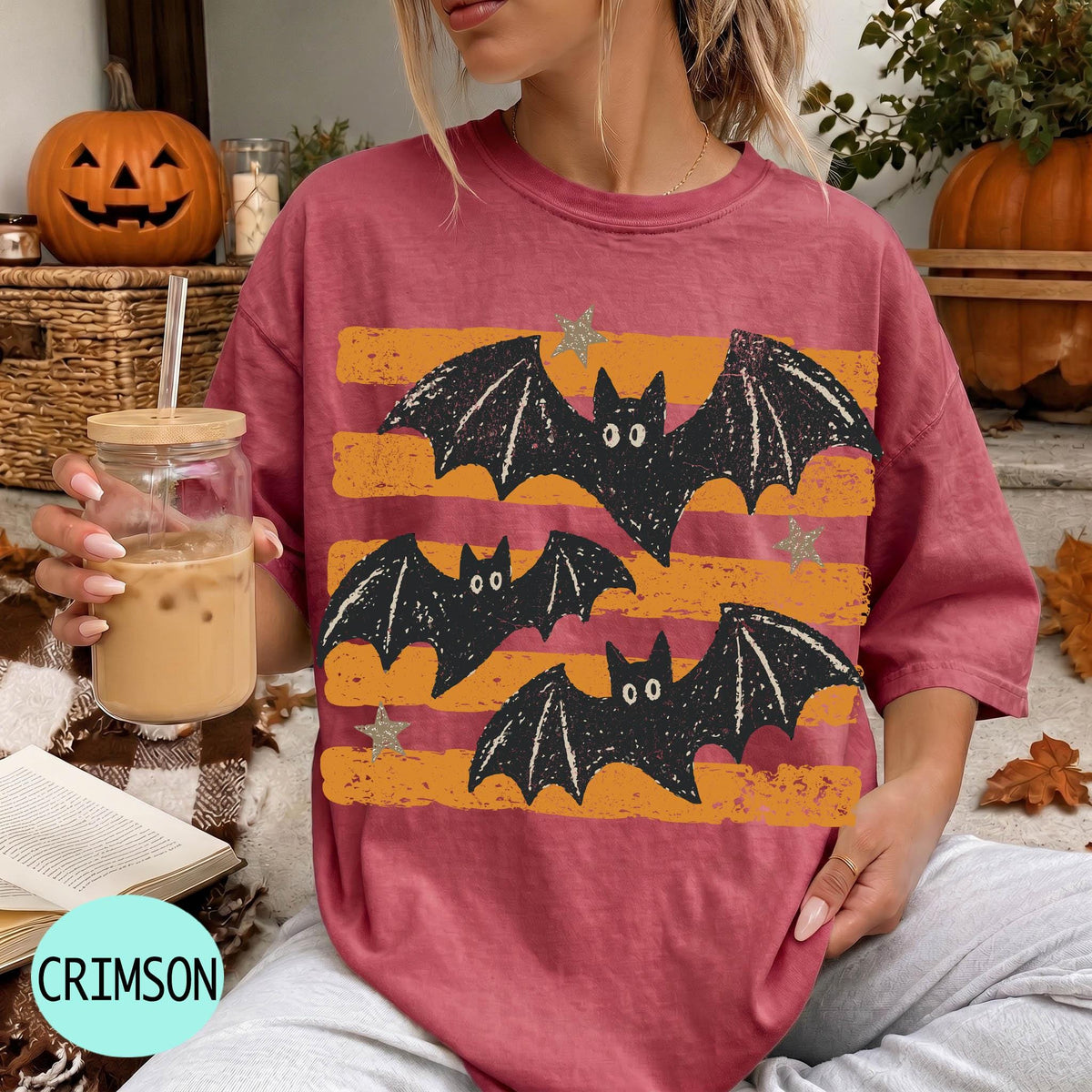 A woman wearing a red t-shirt with a bat design is sitting on a couch, holding a drink in a glass. There are pumpkins and a pumpkin decoration in the background, suggesting a Halloween theme.