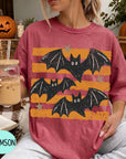 A woman wearing a red t-shirt with a bat design is sitting on a couch, holding a drink in a glass. There are pumpkins and a pumpkin decoration in the background, suggesting a Halloween theme.