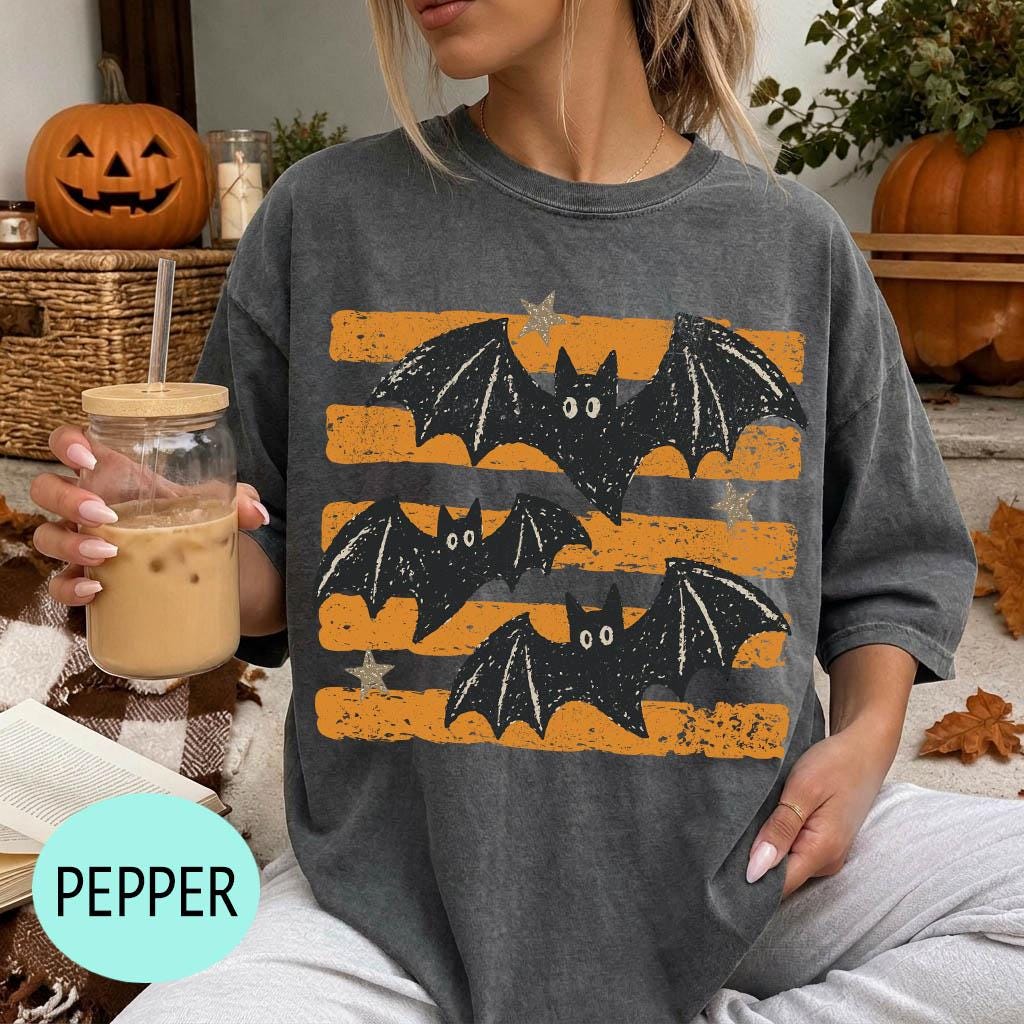 A woman wearing a gray t-shirt with a graphic of bats and stars is sitting on a couch, holding a drink in a glass.