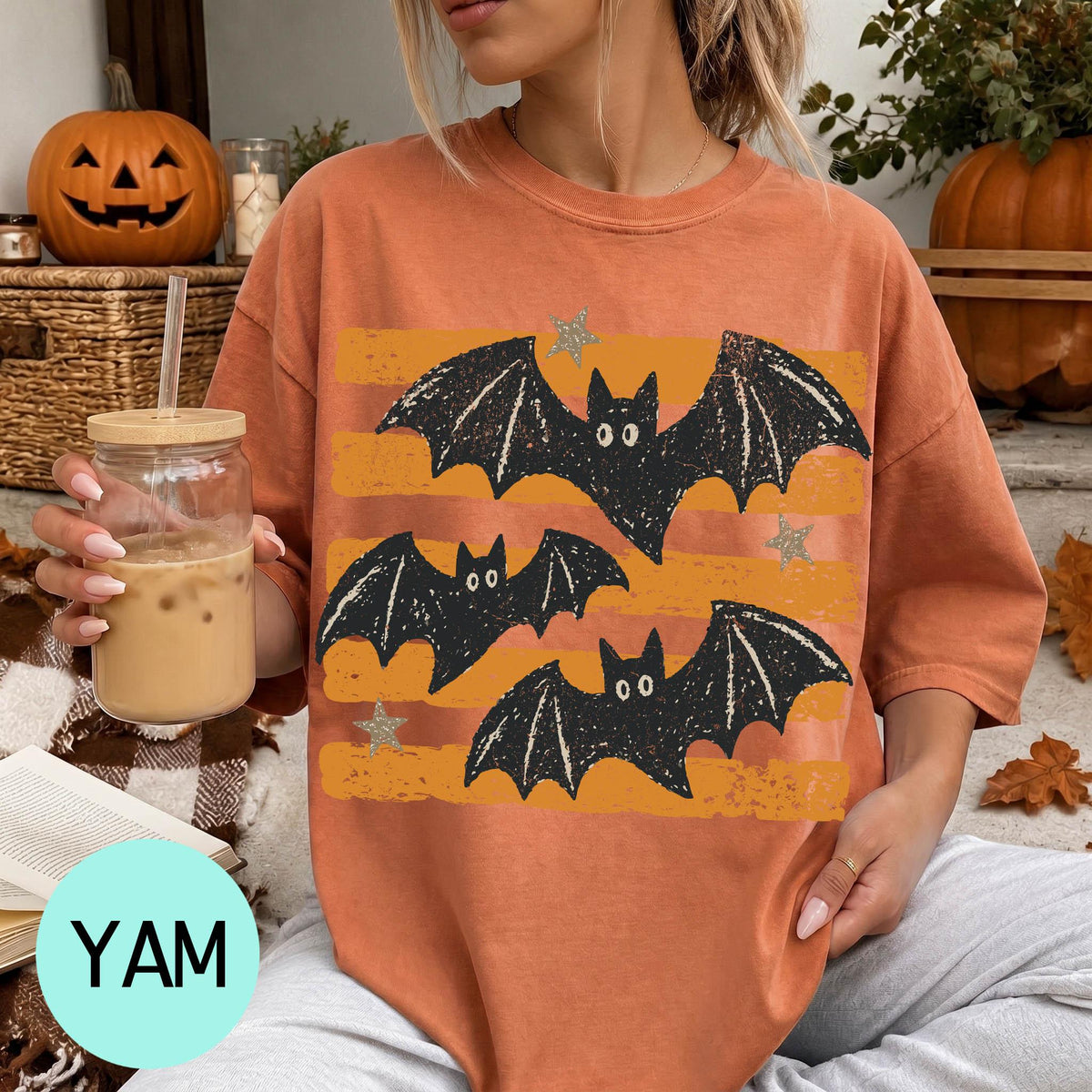 A woman wearing a t-shirt with a bat design is sitting on a couch, holding a drink in a glass. There are pumpkins and a pumpkin decoration in the background, suggesting a Halloween theme.